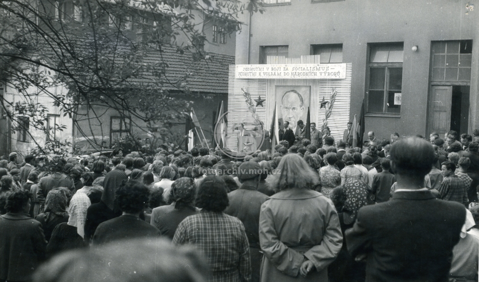 Fotografie - Předvolební shromáždění na ulici - nápis "Jednotni v boji za socialismus - jednotni k volbám do národních výborů" a portrét A. Zápotockého